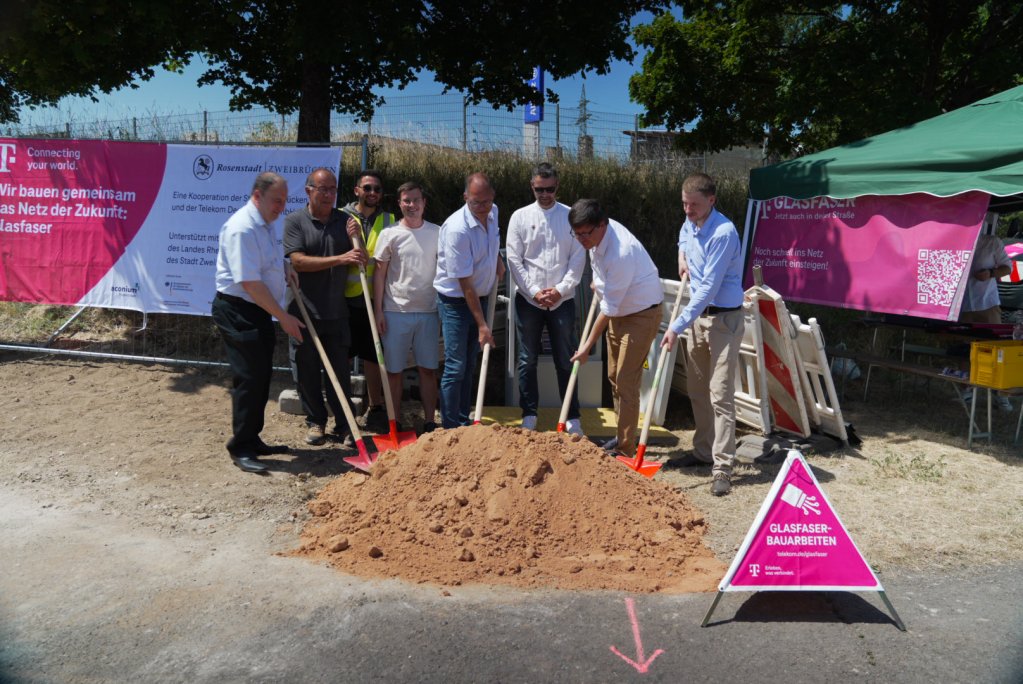Gemeinsamer Spatenstich in Zweibrücken. Auf dem Bild sind Personen der Stadt, Telekom und den Fördergeldgebern zu sehen. Die Personen haben eine Schaufel in der Hand und werfen den Sand damit in die Luft. Im Hintergrund ist ein Banner zu sehen mit den Logos von Telekom, Stadt, aconium und Land Rheinland-Pfalz