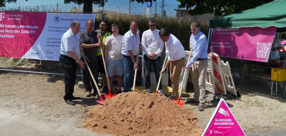 Gemeinsamer Spatenstich in Zweibrücken. Auf dem Bild sind Personen der Stadt, Telekom und den Fördergeldgebern zu sehen. Die Personen haben eine Schaufel in der Hand und werfen den Sand damit in die Luft. Im Hintergrund ist ein Banner zu sehen mit den Logos von Telekom, Stadt, aconium und Land Rheinland-Pfalz